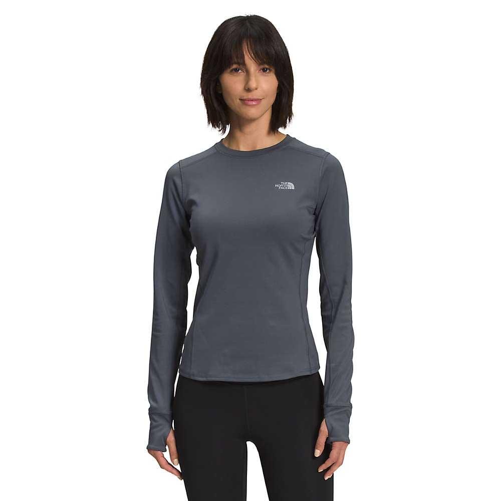 A woman with dark hair wears The North Face Womens Winter Warm Essential Crew, black pants, and an adjustable suspension backpack as she stands facing forward and smiles against a plain white background.