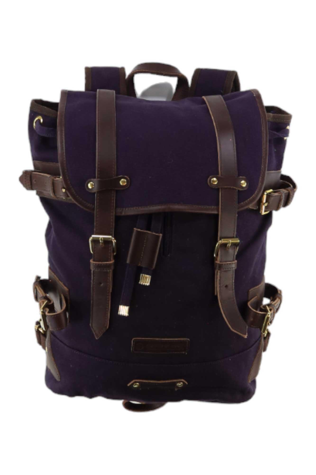 The United By Blue Derby Tier Backpack is a dark purple canvas travel bag with brown leather straps, brass buckles, flap top, side pockets, and top handle—perfect for stylish adventures.