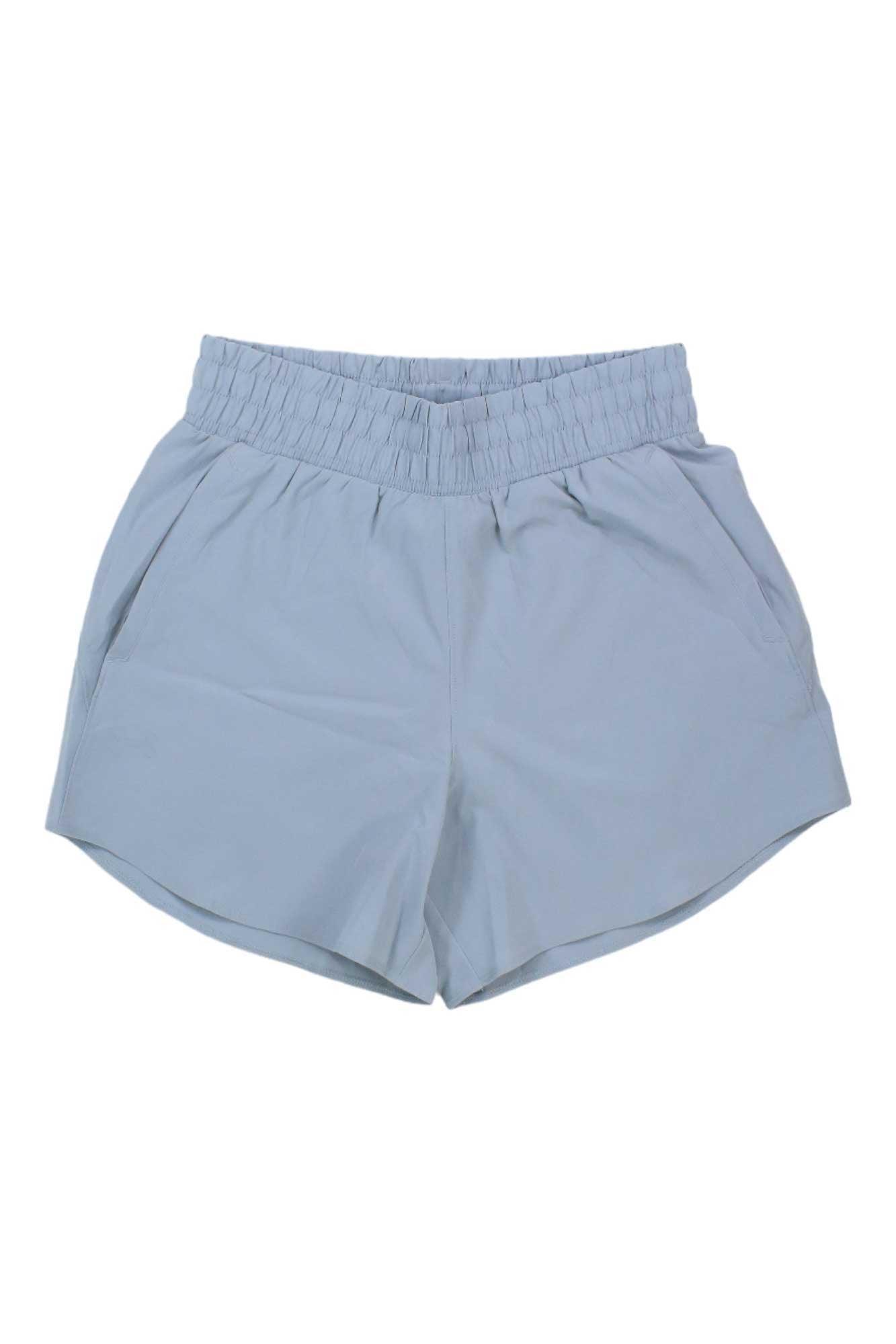 The Under Armour Women's Flex Woven 5 Inch Short features light blue 4-way stretch fabric, an elastic waistband, and side pockets for comfort and flexibility. Shown laid flat on a white background.