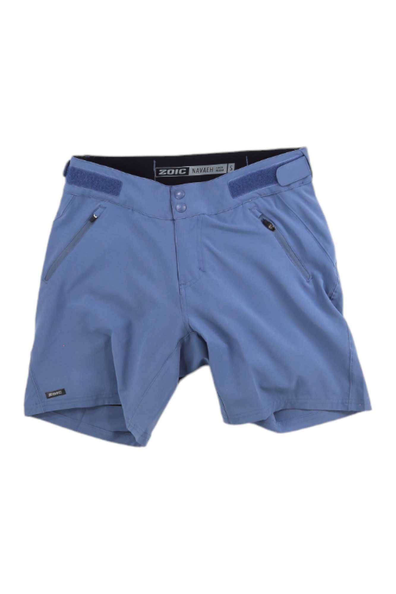 The Zoic Women's Navaeh 7 Short - Essential Liner is a light blue mid-rise casual short featuring zippered side pockets, adjustable waistband tabs, a button closure, and packable polyester material. Shown laid flat on a white background.