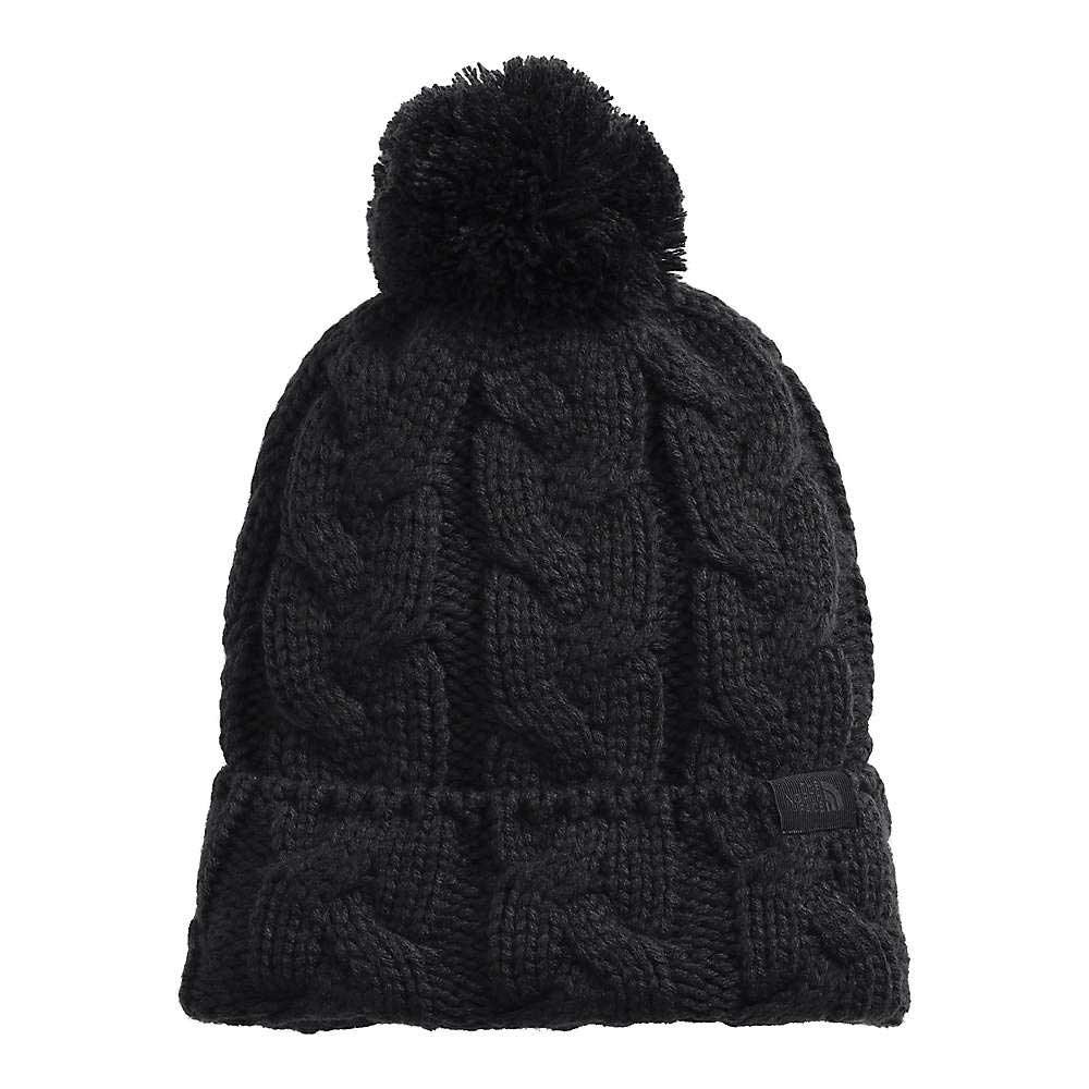 The North Face Cable Minna Pom Beanie is a black, thick cable knit beanie with a large pom-pom and folded brim, made from recycled polyester fleece. It features a small rectangular fabric tag on the brim.