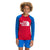 A smiling child with curly hair wears The North Face Boys Amphibious LS Sun Tee, a red and blue long-sleeve swim shirt with UPF 40+ protection and blue shorts, standing against a plain white background.