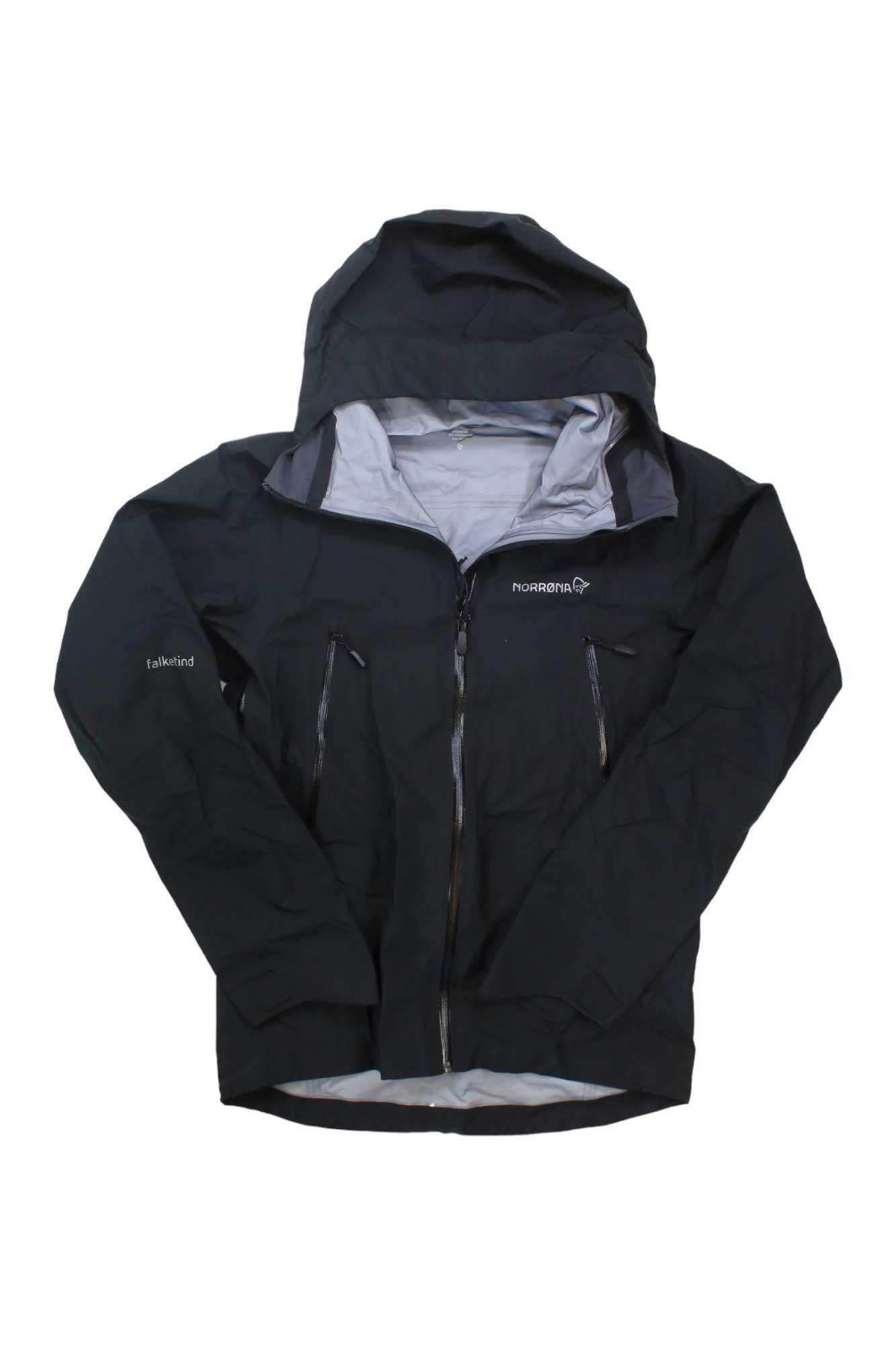 The Norrona Mens Falketind GTX Jacket is a black Gore-Tex hiking jacket with a light gray lining, zippered chest pockets, and Norrona and Falketind logos on the front and sleeve.