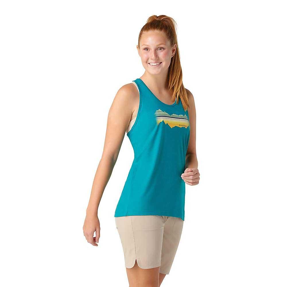 A woman with long red hair in a ponytail models the Smartwool Women's Mountain Horizon Graphic Tank and beige shorts, smiling and posing against a plain white background.