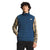 A man wears The North Face Mens Belleview Stretch Down Vest in blue over a yellow plaid shirt and black pants, standing facing forward with one hand in his pocket. The vest’s brand logo is clearly visible.