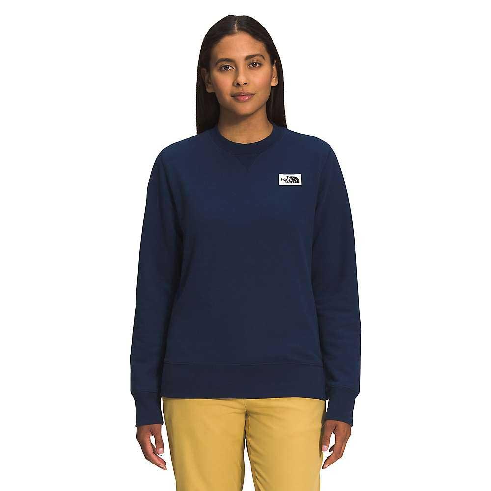A woman with long, straight dark hair wears The North Face Womens Heritage Patch Crew in navy blue and yellow pants, standing facing forward against a plain white background.