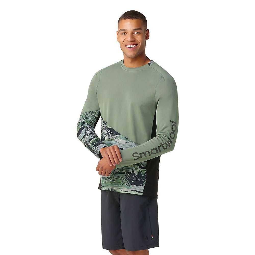 A man wearing the Smartwool Mens Mountain Bike LS Jersey, a long-sleeve green patterned top made from moisture-wicking merino wool, and dark shorts stands smiling with arms crossed in front of a white background.