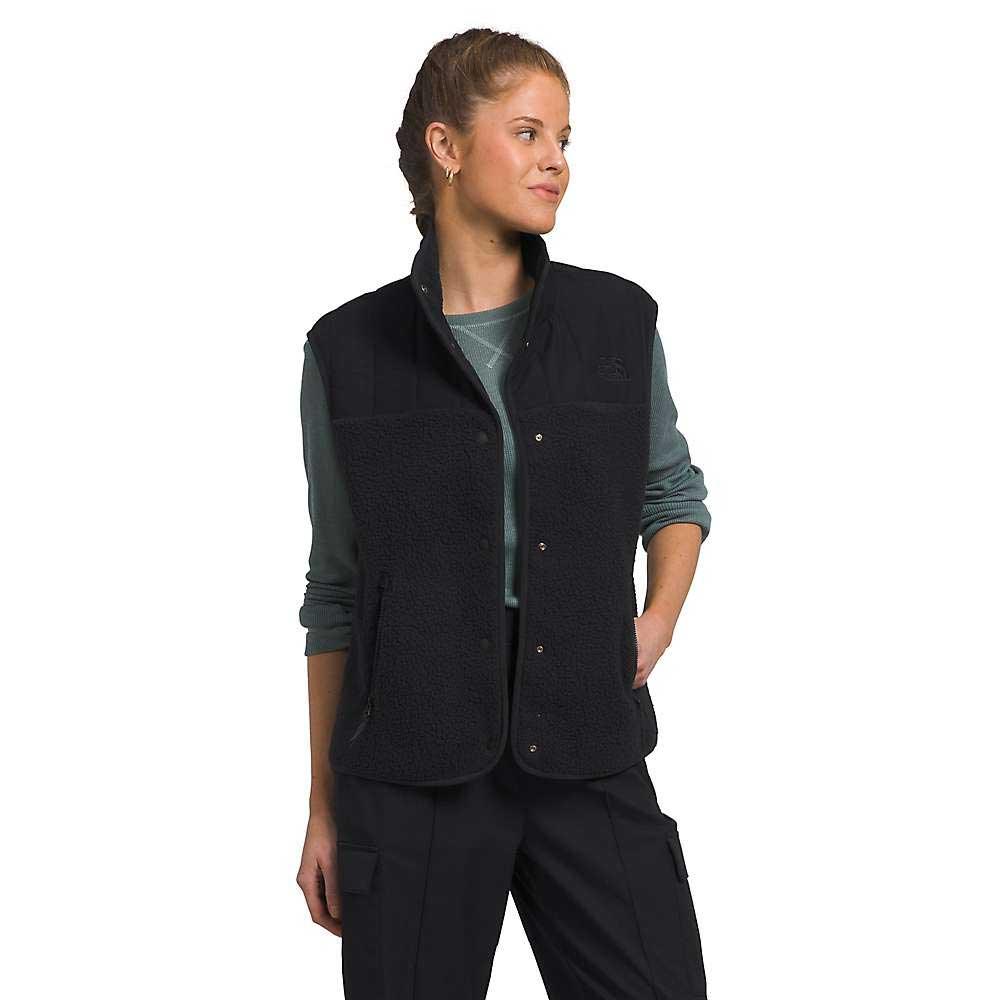 A woman wears The North Face Women's Cragmont Fleece Vest in black over a long-sleeve green shirt and black pants, standing with one hand in her pocket, looking to the side and smiling slightly.