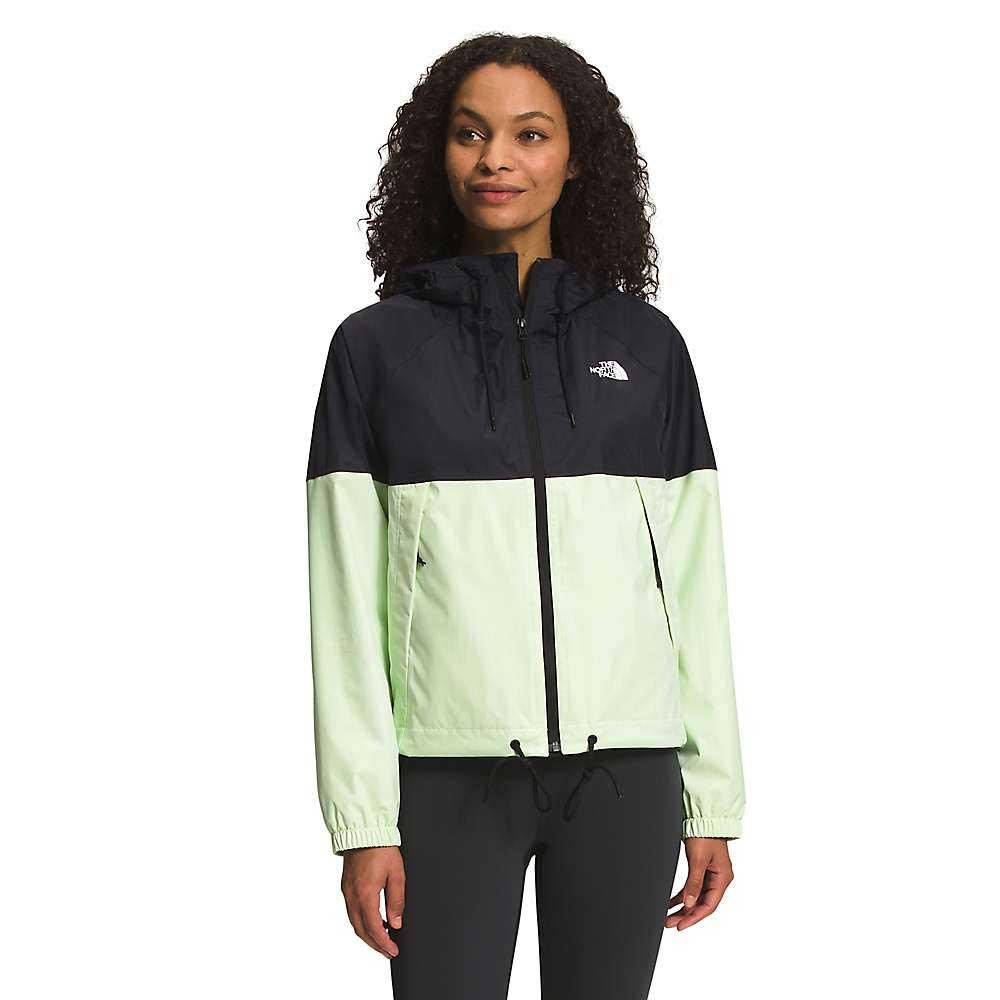 A woman with curly hair wears the black and light green The North Face Womens Antora Rain Hoodie, featuring an adjustable hood, front zipper, and elastic cuffs. She pairs it with black pants against a plain white background.