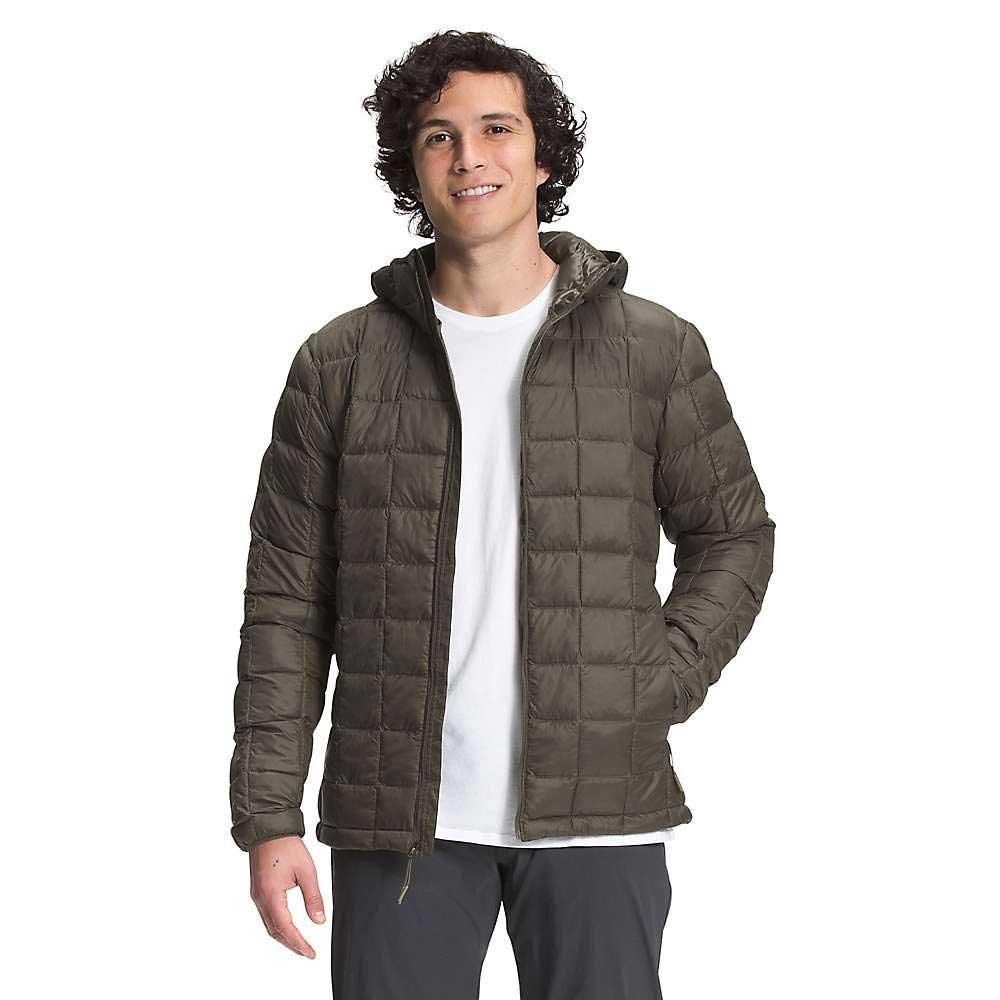 A person with curly dark hair wears The North Face Mens ThermoBall Eco Hoodie, a PFAS-free puffer jacket with a hood and zippered front, over a white t-shirt and dark pants. Smiling at the camera, they stand with one hand in a pocket.