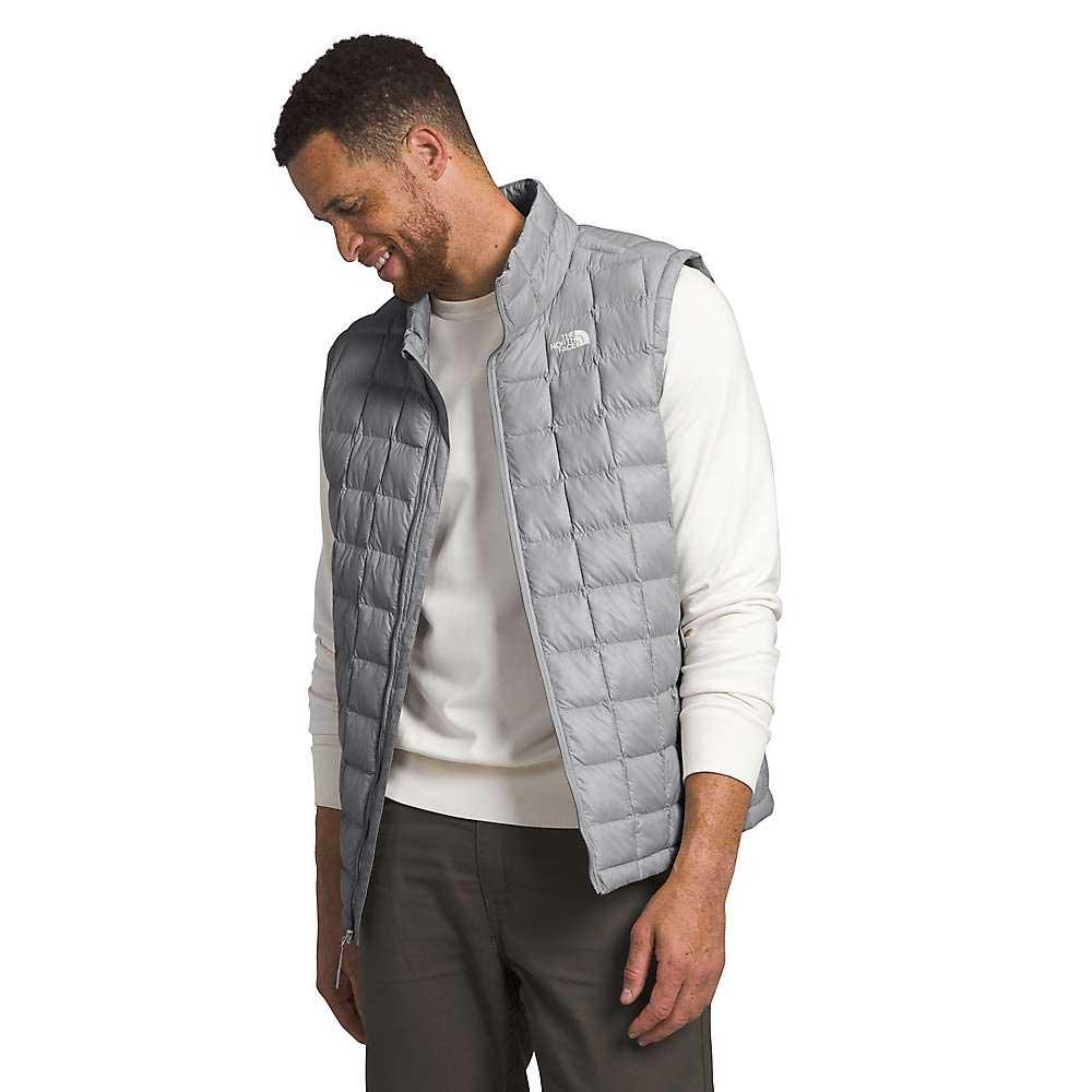 A man wearing The North Face Men's Big ThermoBall Eco 2.0 Vest in light gray over a white long-sleeve shirt and gray pants stands, smiling as he looks down with his left hand in his pocket.
