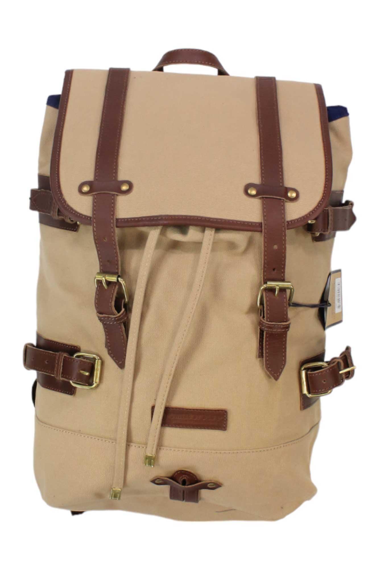 The United By Blue Derby Tier Backpack is a tan canvas bag with brown leather straps, gold buckles, a drawstring closure, top flap, side straps, and a top handle—perfect for stylish adventures.