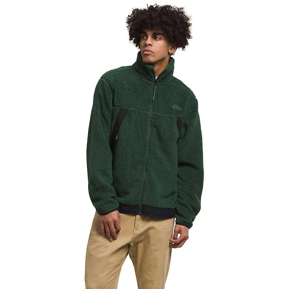 A young man with curly hair wears The North Face Men's Campshire Fleece Jacket in dark green, made from recycled polyester fleece, and beige pants, standing with one hand in his pocket against a plain white background.