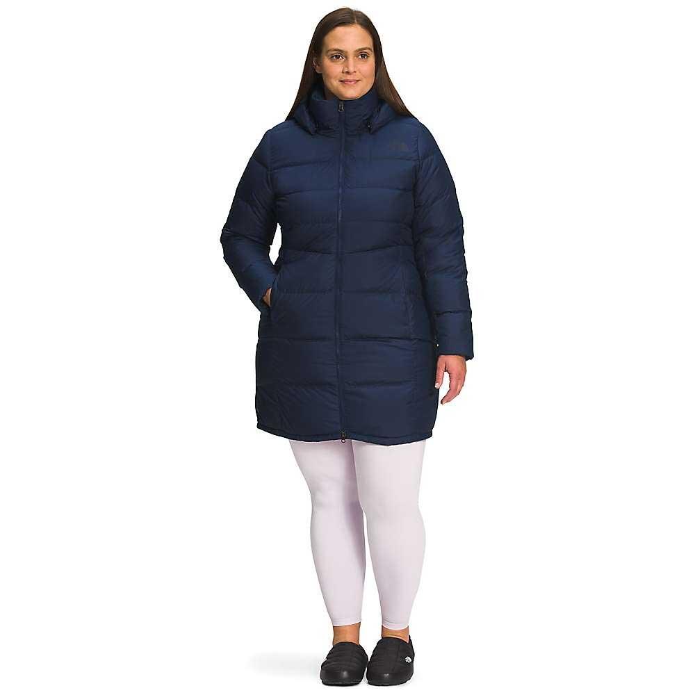 A woman wearing The North Face Women's Plus Metropolis Parka in dark blue, with a water-repellent finish and goose down insulation, stands smiling at the camera, hands in pockets, white leggings, and black slip-on shoes against a white background.