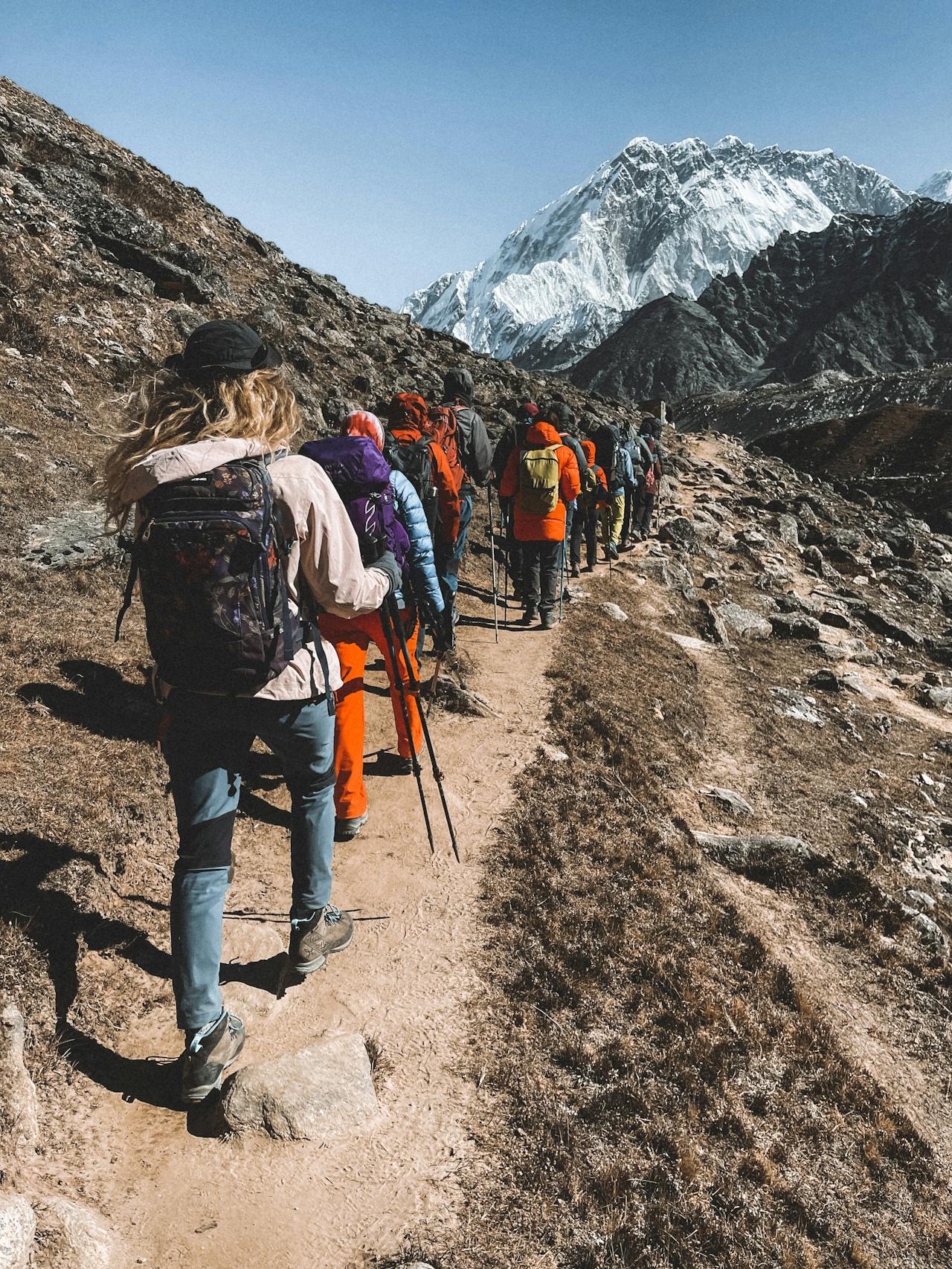 A row of people, hiking in the mountains with snow capped peaks in the background