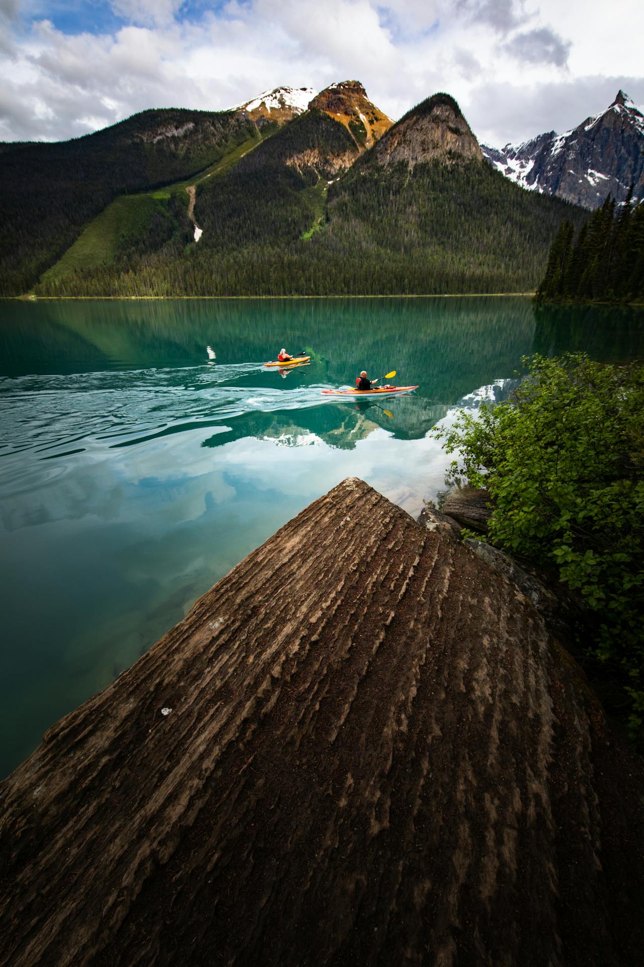 Two people in kayaks, paddling across a turquoise lake with mountains in the background
