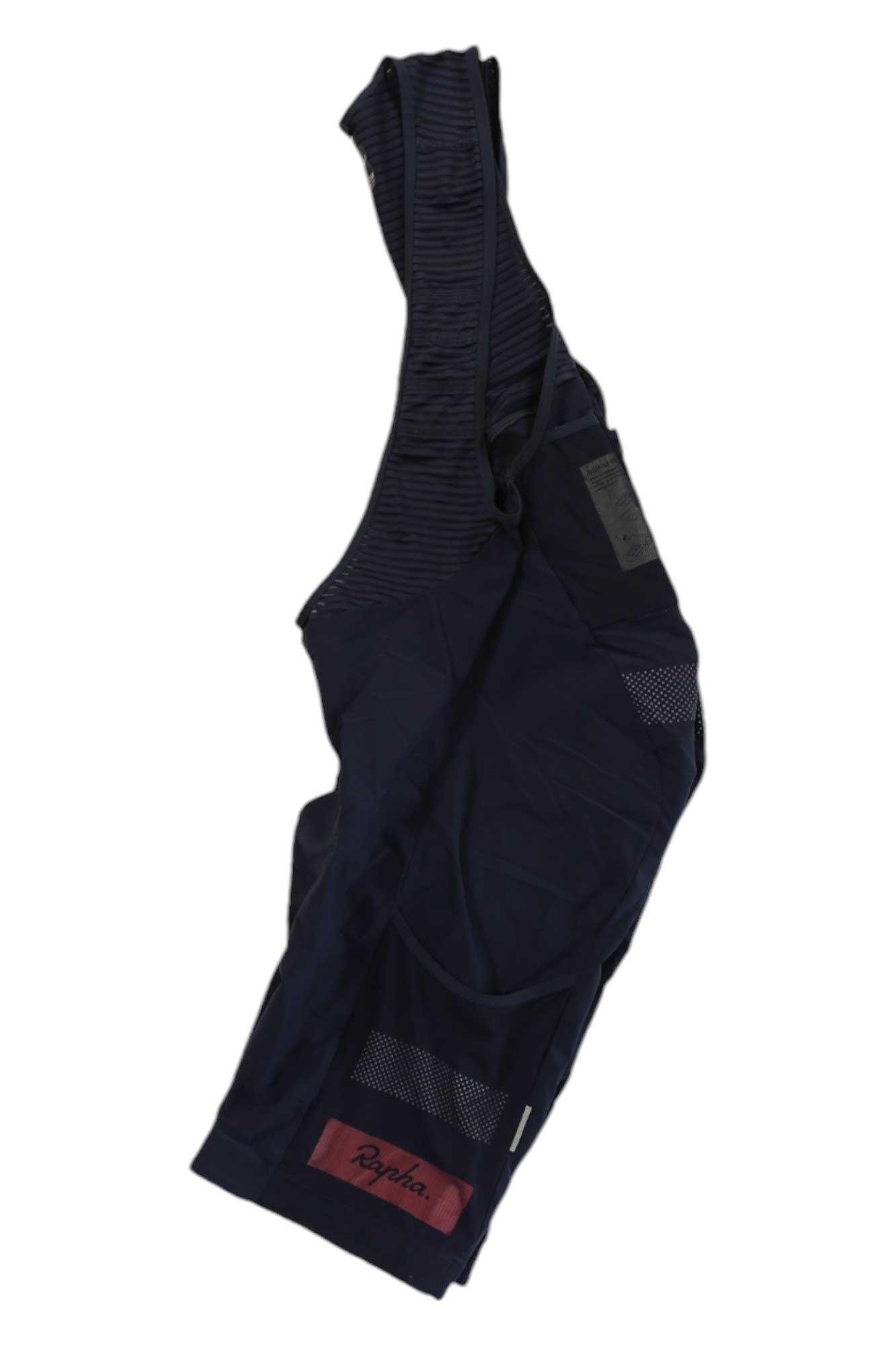 A pair of dark navy blue Rapha Cargo Bib Shorts, featuring mesh straps and a maroon logo tag on the lower leg, made from dense-knit fabric for cycling comfort, is laid flat on a white background.